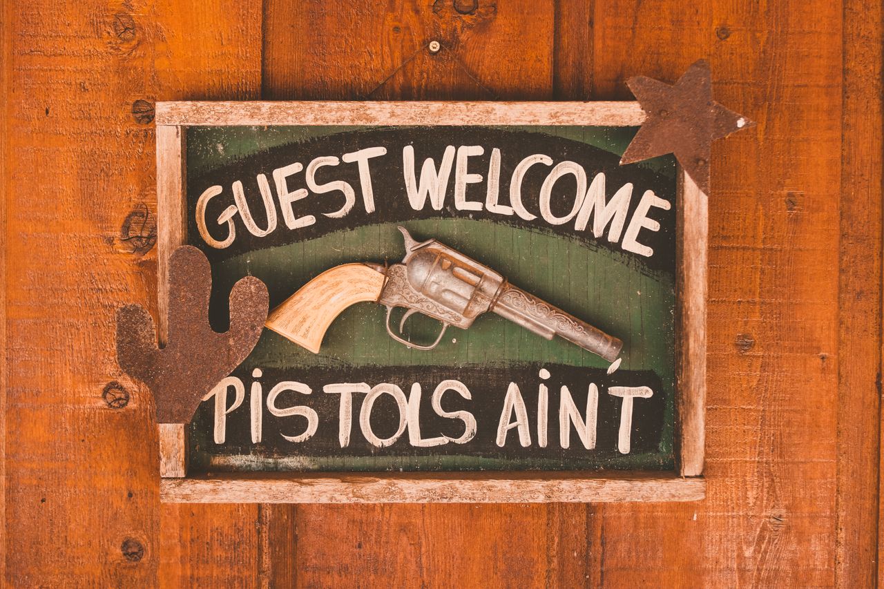 Framed rectangular signboard with inscription Guest Welcome Pistols Aint decorated with artificial gun and silhouettes of cactus and star on wooden door